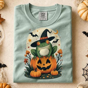 May include: A light green t-shirt featuring a graphic of a green frog wearing a black witch hat, sitting atop a large orange jack-o'-lantern. The design includes small orange mushrooms, bats, and stars against a pale yellow moon background.