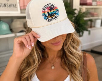 Könnte beinhalten: Eine weiße Trucker-Cap mit Netzrücken und einem Grafikdruck auf der Vorderseite. Das Design zeigt die Silhouette einer Person auf einem Surfbrett unter einer Palme vor einem Sonnenuntergang und den Schriftzug COASTAL COWGIRL in schwarzen Buchstaben.