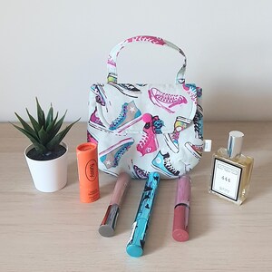 May include: A small, light-coloured handbag with a handle, patterned with colourful high-top trainers. Several lipsticks, a perfume bottle, and a small potted plant are arranged around the bag on a wooden surface.