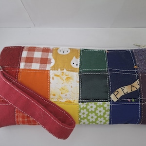 May include: A rectangular zippered pouch made of multicolored fabric patches in a grid pattern. The patches feature various colors including red, orange, yellow, green, blue, and purple, with patterns like gingham, flowers, and cat faces. A red wrist strap is attached.
