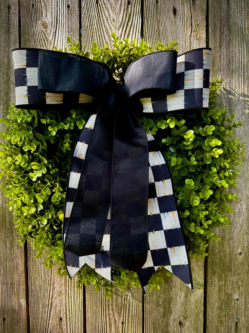 May include: A round green wreath with a large black bow featuring a checkered pattern in black and white. The wreath is set against a weathered wooden background, creating a rustic and elegant aesthetic.