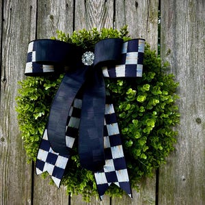 May include: A decorative wreath with a lush green base and a large black bow. The bow features a checkered pattern on the edges and a sparkling jewel in the center. The wreath is displayed against a weathered wooden fence.