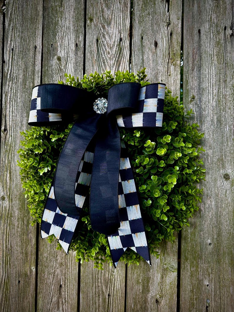 May include: A decorative wreath with a lush green base and a large black bow. The bow features a checkered pattern on the edges and a sparkling jewel in the center. The wreath is displayed against a weathered wooden fence.