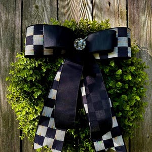 May include: A decorative wreath featuring a lush green base, adorned with a large black bow. The bow has a checkered pattern on the edges and a sparkling jewel in the center. The wreath is displayed against a weathered wooden fence.