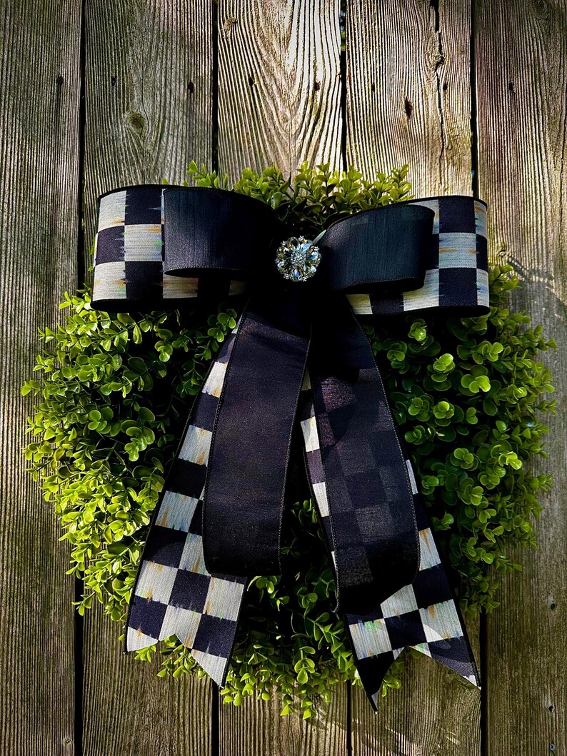 May include: A decorative wreath featuring a lush green base, adorned with a large black bow. The bow has a checkered pattern on the edges and a sparkling jewel in the center. The wreath is displayed against a weathered wooden fence.