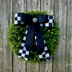 May include: A round green wreath with a large black bow featuring a checkered pattern. The bow is adorned with a sparkling, round jewel. The wreath is hanging on a weathered wooden fence.