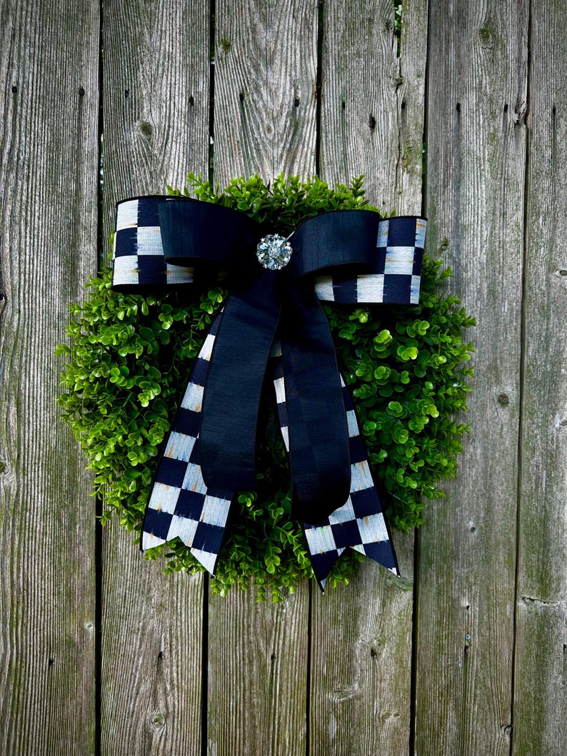 May include: A round green wreath with a large black bow featuring a checkered pattern. The bow is adorned with a sparkling, round jewel. The wreath is hanging on a weathered wooden fence.