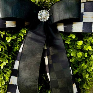 May include: A decorative wreath featuring a lush green base, a large black bow, and a sparkling rhinestone embellishment. The bow has a checkered pattern on the edges. The wreath is suitable for home decor.