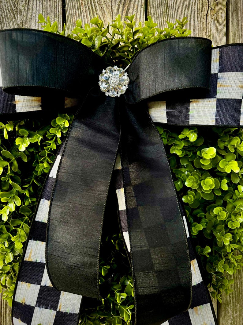 May include: A decorative wreath featuring a lush green base, a large black bow, and a sparkling rhinestone embellishment. The bow has a checkered pattern on the edges. The wreath is suitable for home decor.