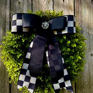 May include: A decorative wreath featuring a lush green base, adorned with a large black bow. The bow has a checkered pattern on the edges and a sparkling jewel in the center. The wreath is displayed against a weathered wooden fence.