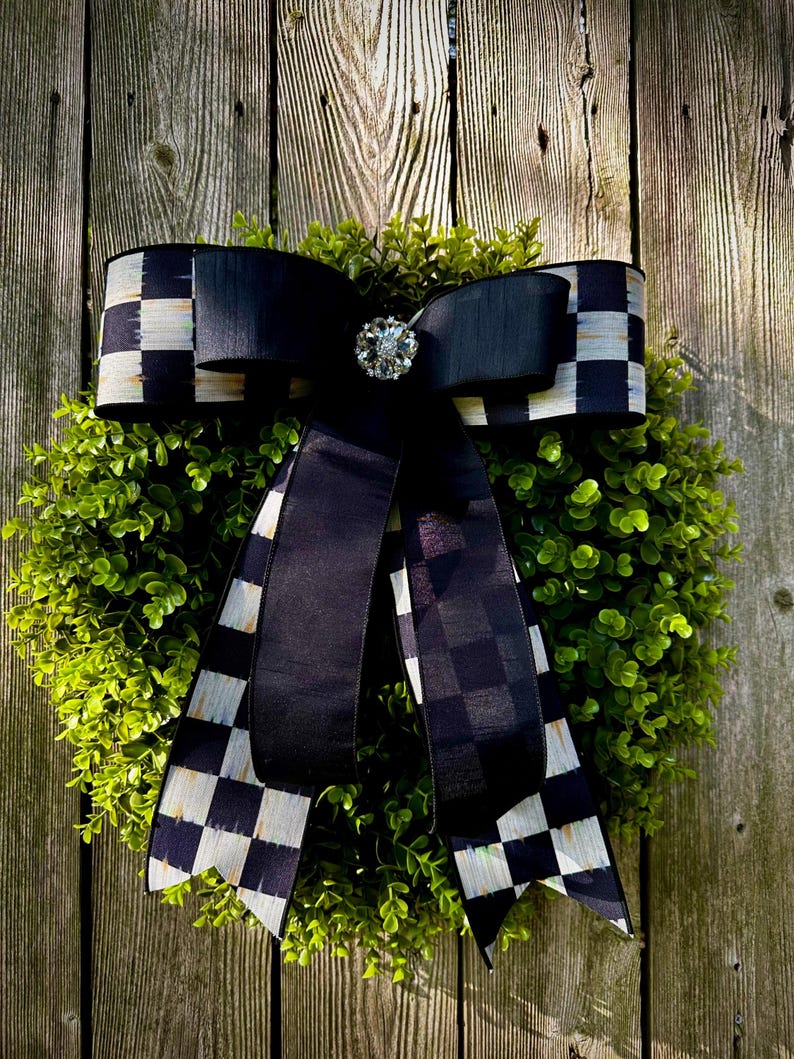 May include: A decorative wreath featuring a lush green base, adorned with a large black bow. The bow has a checkered pattern on the edges and a sparkling jewel in the center. The wreath is displayed against a weathered wooden fence.