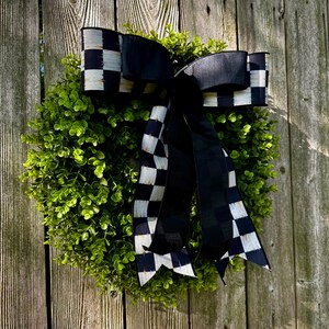 May include: A round, green wreath with a large black and white checkered bow. The bow's ribbon has a bold pattern, and the wreath is made of small, leafy green foliage. The background is a weathered wooden fence.