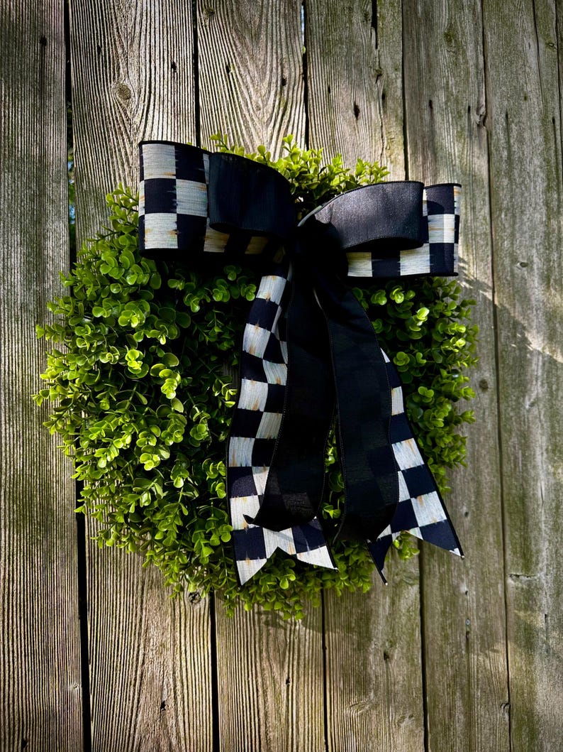 May include: A round, green wreath with a large black and white checkered bow. The bow's ribbon has a bold pattern, and the wreath is made of small, leafy green foliage. The background is a weathered wooden fence.