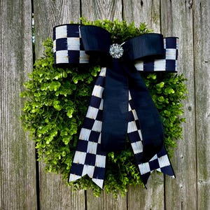 May include: A decorative wreath featuring a lush green base, adorned with a large black and white checkered bow. A sparkling jewel embellishment sits at the bow's center. The wreath is displayed against a weathered wooden fence.