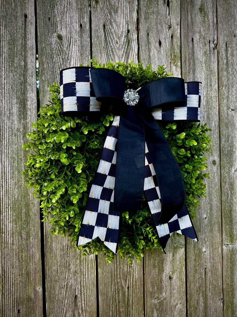May include: A decorative wreath featuring a lush green base, adorned with a large black and white checkered bow. A sparkling jewel embellishment sits at the bow's center. The wreath is displayed against a weathered wooden fence.