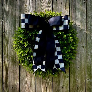 May include: A round, green wreath with a large black and white checkered bow. The wreath is made of small, leafy green plants and is hanging on a weathered wooden fence. The bow adds a decorative touch.