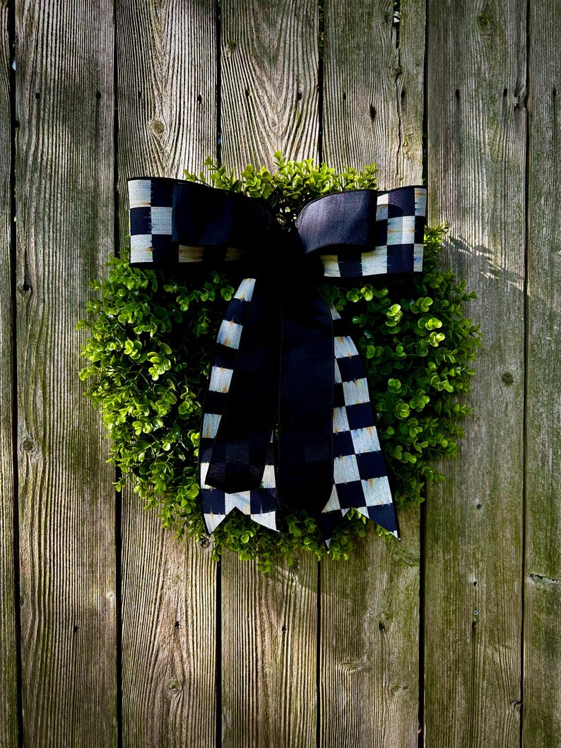 May include: A round, green wreath with a large black and white checkered bow. The wreath is made of small, leafy green plants and is hanging on a weathered wooden fence. The bow adds a decorative touch.