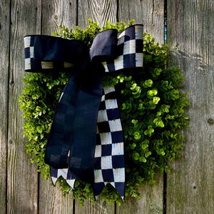 May include: A round, green wreath with a large black and white checkered bow. The wreath is made of small, leafy green plants and is set against a weathered wooden fence.
