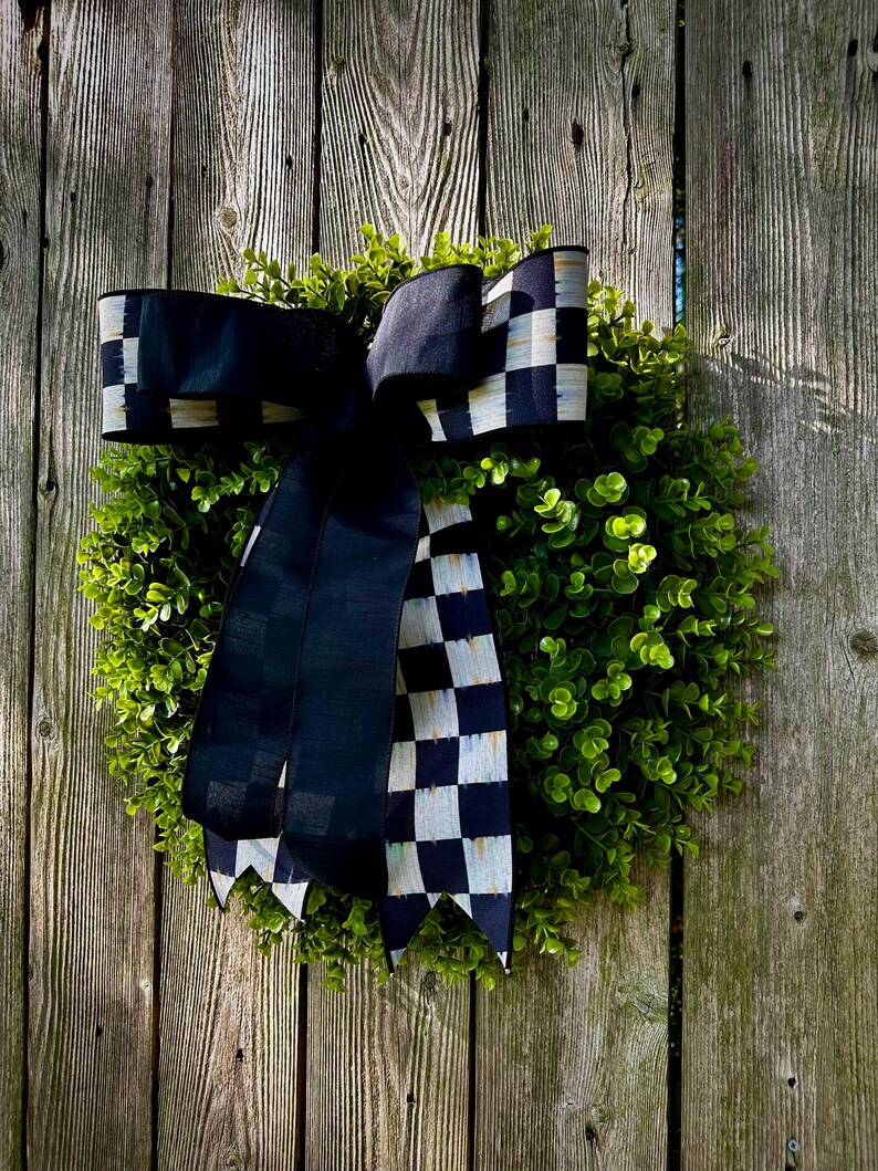 May include: A round, green wreath with a large black and white checkered bow. The wreath is made of small, leafy green plants and is set against a weathered wooden fence.