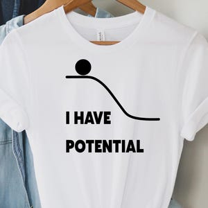 Könnte beinhalten: Ein weißes kurzärmeliges T-Shirt mit einer schwarzen Grafik, die einen Ball auf einer geschwungenen Linie zeigt, sowie dem Text I HAVE POTENTIAL. Das T-Shirt hängt an einem Holzkleiderbügel, im Hintergrund sind ein blaues Jeanshemd und ein brauner Hut.
