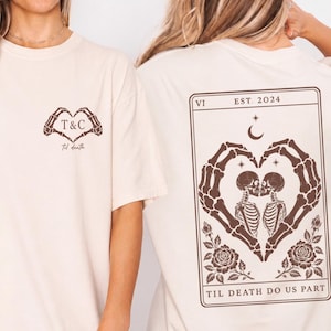 Pode incluir: Uma t-shirt cor de creme com uma impressão gráfica castanha. A frente exibe mãos de esqueleto a formar um coração em torno do texto T&C e Til death. As costas mostram um design de carta de tarot com esqueletos a beijar-se, rosas e o texto VI EST. 2024 TIL DEATH DO US PART.