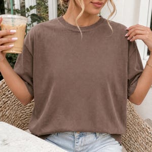 May include: A solid brown short-sleeve t-shirt with a crew neckline. The garment features a relaxed fit and a smooth, matte fabric texture. The shirt is worn with light blue denim jeans, creating a simple and neutral appearance for daily wear.