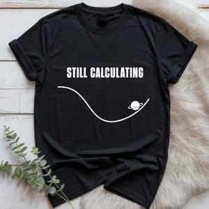 Op de afbeelding: Een zwart T-shirt met korte mouwen voorzien van de witte tekst STILL CALCULATING boven een witte afbeelding van een gebogen lijn en een kleine planeet met ringen. Het shirt heeft een ronde hals en ligt plat uitgevouwen.