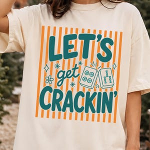 May include: A cream-coloured short-sleeve t-shirt featuring a graphic print with vertical orange stripes. The text reads "LET'S get CRACKIN'" in dark teal letters, accompanied by illustrations of two mahjong tiles, small stars, and flowers.