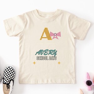 May include: A cream-coloured short-sleeve t-shirt featuring a large yellow letter A with black polka dots and a pink bow. Below, the text reads AVERY in teal and SCHOOL DAYS in black, accented by two small yellow four-pointed star icons.