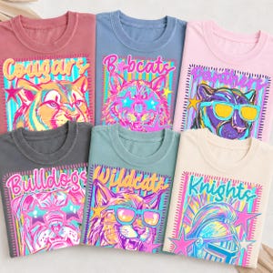 May include: A collection of six crew neck t-shirts in various colors including dusty rose, blue, light pink, charcoal, sage green, and cream. Each shirt features a vibrant graphic print of an animal or knight wearing star-shaped sunglasses.