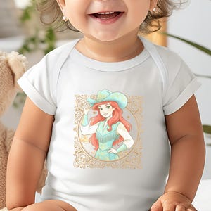 K&ouml;nnte beinhalten: Wei&szlig;er Baby-Body mit einer Cartoon-Illustration einer Cowgirl mit roten Haaren und blauem Hut. Das Design ist von einem goldenen Zierrahmen umgeben. Das Wort "WHITE" ist unten aufgedruckt.