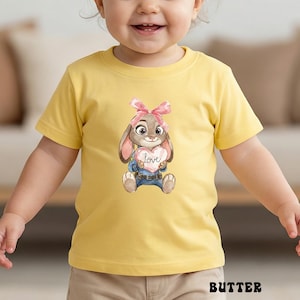 Peut inclure: T-shirt jaune &agrave; manches courtes avec un lapin de dessin anim&eacute; tenant un c&oelig;ur avec le mot "love". Le lapin porte un n&oelig;ud rose et un jean bleu. Le mot "BUTTER" est imprim&eacute; en bas.