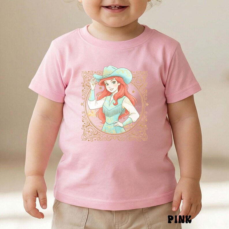 K&ouml;nnte beinhalten: Rosa kurz&auml;rmeliges T-Shirt mit einer Grafik einer Cowgirl mit roten Haaren und einem blauen Hut. Die Cowgirl befindet sich in einem goldfarbenen, verzierten Rahmen. Das Wort "PINK" ist unten auf dem Shirt in Schwarz aufgedruckt.