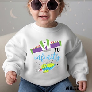 May include: White sweatshirt with a colorful graphic featuring a rocket ship and the phrase "TO infinity" in blue. The design includes green, purple, and red accents. The word "WHITE" is printed at the bottom.