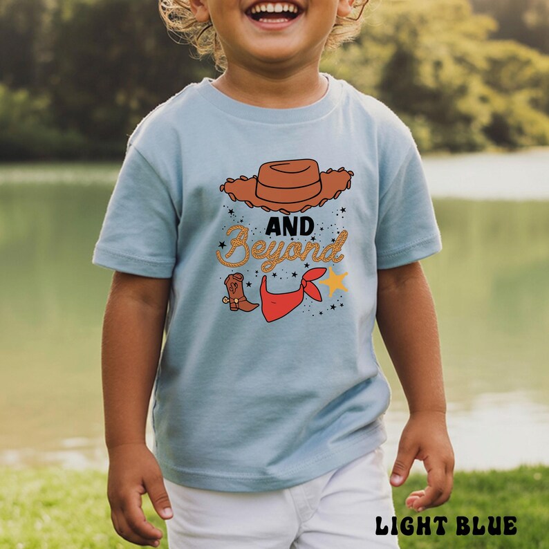 May include: Light blue t-shirt with a cowboy-themed graphic. The design includes a brown cowboy hat, a red bandana, a cowboy boot, and the words "AND Beyond" in a decorative font. The words "LIGHT BLUE" are at the bottom.
