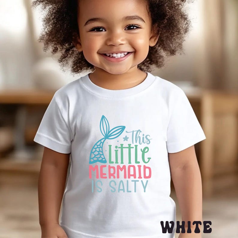 Puede incluir: Camiseta blanca con un dise&ntilde;o de cola de sirena y el texto "This Little Mermaid Is Salty" en azul, verde y rosa. La palabra "WHITE" est&aacute; en la parte inferior. La camiseta la lleva un ni&ntilde;o peque&ntilde;o.