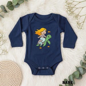 Puede incluir: Body de beb&eacute; azul marino con mangas largas. La parte delantera presenta una sirena de dibujos animados con cabello rubio montada en una tortuga verde, rodeada de peces naranjas. La palabra "NAVY" est&aacute; impresa en la parte inferior derecha.