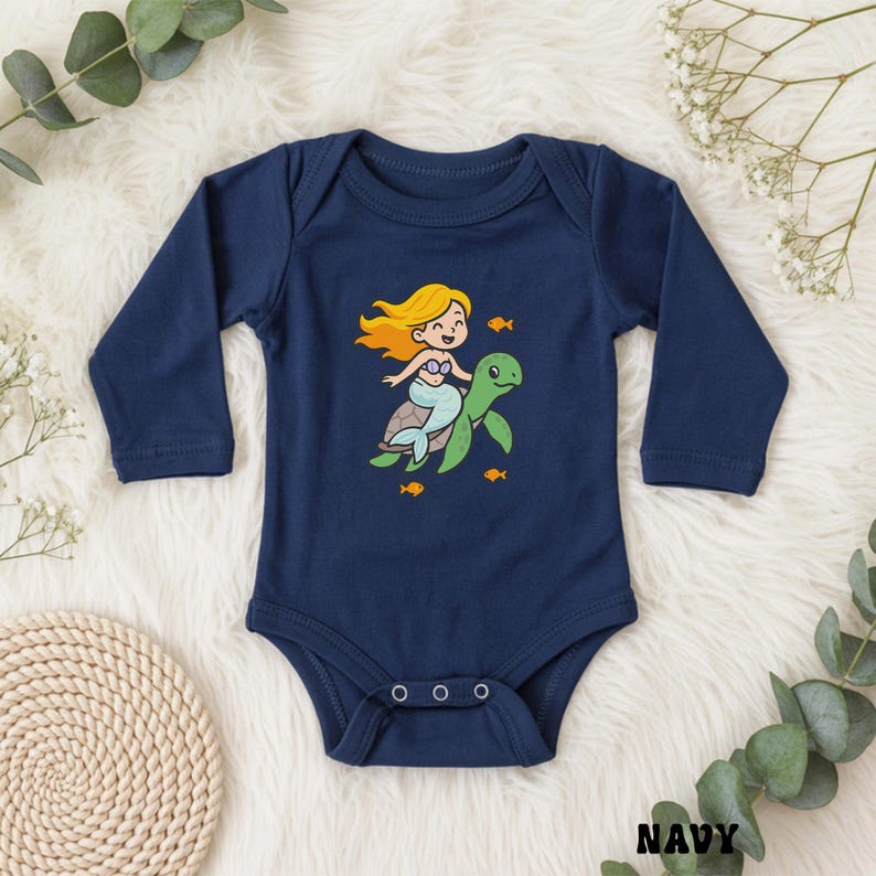 Puede incluir: Body de beb&eacute; azul marino con mangas largas. La parte delantera presenta una sirena de dibujos animados con cabello rubio montada en una tortuga verde, rodeada de peces naranjas. La palabra "NAVY" est&aacute; impresa en la parte inferior derecha.