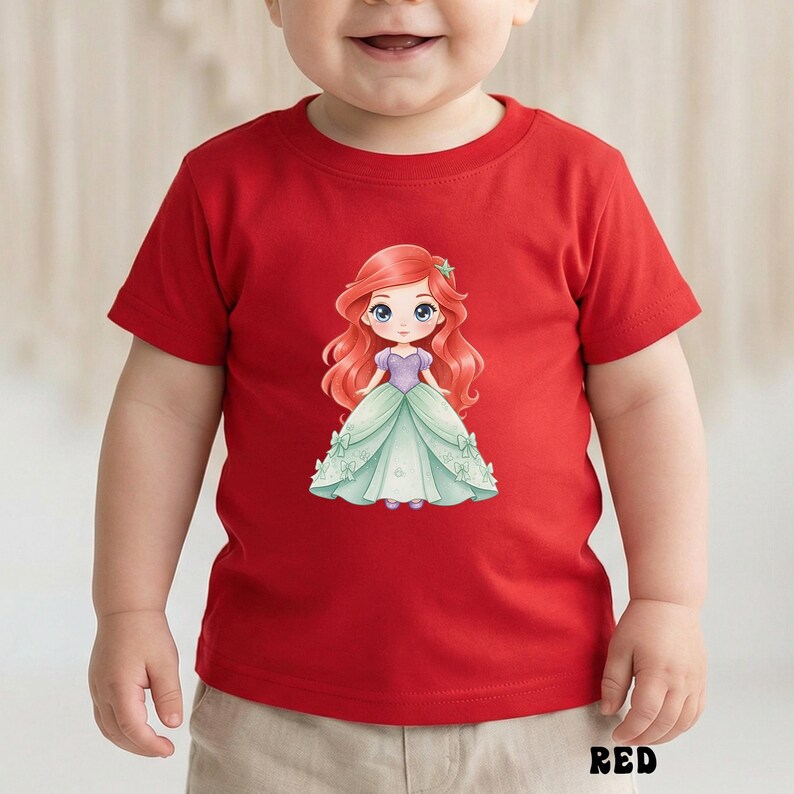 Puede incluir: Una camiseta roja de manga corta con un gr&aacute;fico central de un personaje de dibujos animados con largo cabello rojo ondulado, un corpi&ntilde;o morado y una falda de vestido de gala verde claro adornada con peque&ntilde;os lazos. La palabra RED aparece abajo.
