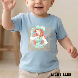 K&ouml;nnte beinhalten: Hellblaues Kleinkind-T-Shirt mit einer Cartoon-Grafik eines rothaarigen Charakters mit Cowboyhut und Fransenweste. Die Grafik ist von einem goldenen, verzierten Rahmen umgeben. Der Text "LIGHT BLUE" steht unten.