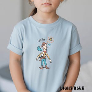 May include: Light blue t-shirt featuring a cartoon illustration of Woody from Toy Story. The design includes the word "WOODY" above the character, a sun, and a cloud. The shirt is labeled "LIGHT BLUE" at the bottom.