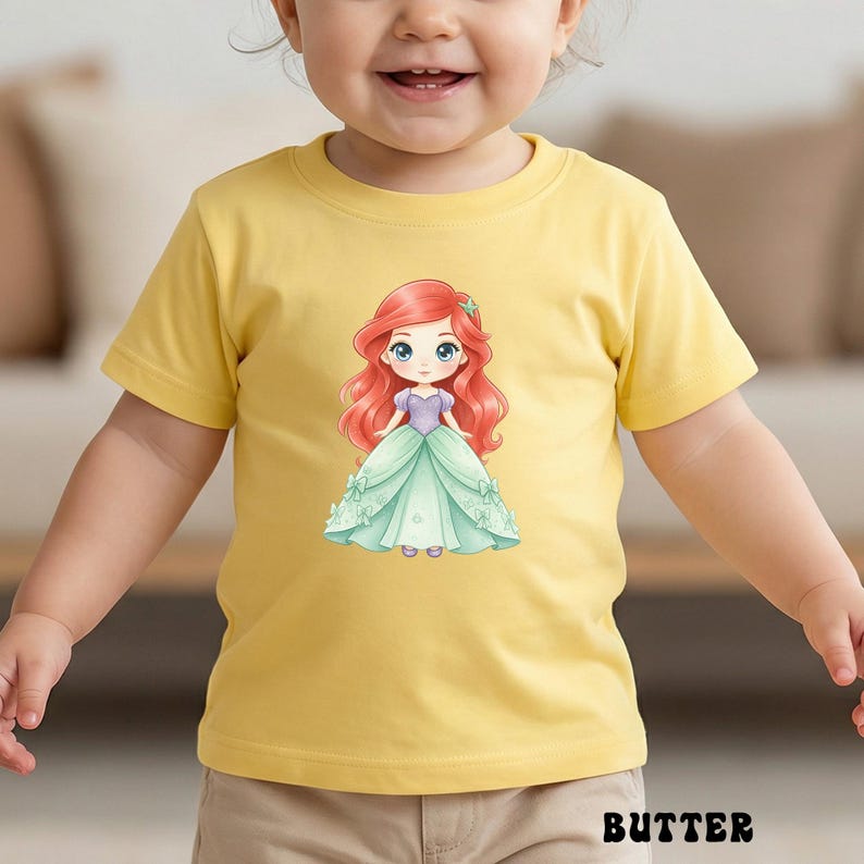 Puede incluir: Una camiseta amarilla de manga corta con una ilustraci&oacute;n gr&aacute;fica central de un personaje de dibujos animados con el pelo largo y rojo, que lleva un corpi&ntilde;o morado y un vestido de gala verde claro con peque&ntilde;os lazos. La palabra BUTTER est&aacute; en negro.
