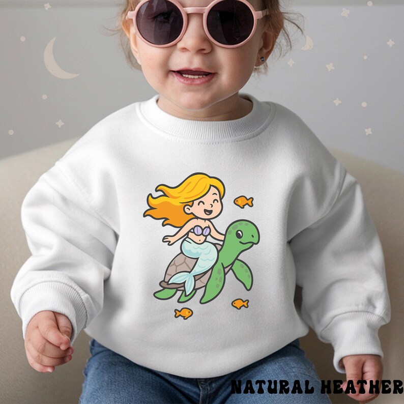 Puede incluir: Sudadera blanca con una sirena de dibujos animados montada en una tortuga verde, rodeada de peque&ntilde;os peces naranjas. La sirena tiene el pelo rubio y una cola azul claro. El texto "NATURAL HEATHER" est&aacute; impreso en la parte inferior.