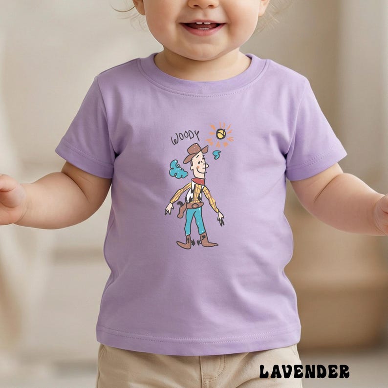 May include: A lavender short-sleeved t-shirt featuring a cartoon illustration of Woody from Toy Story. The illustration includes the word "Woody" above the character. The shirt is made of a soft material.
