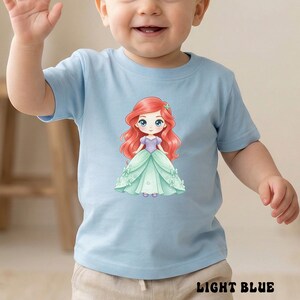 Puede incluir: Una camiseta de manga corta de color azul claro con un estampado gr&aacute;fico de un personaje de dibujos animados con pelo largo rojo, un corpi&ntilde;o morado y un vestido verde claro. El personaje tiene grandes ojos azules y un accesorio verde.