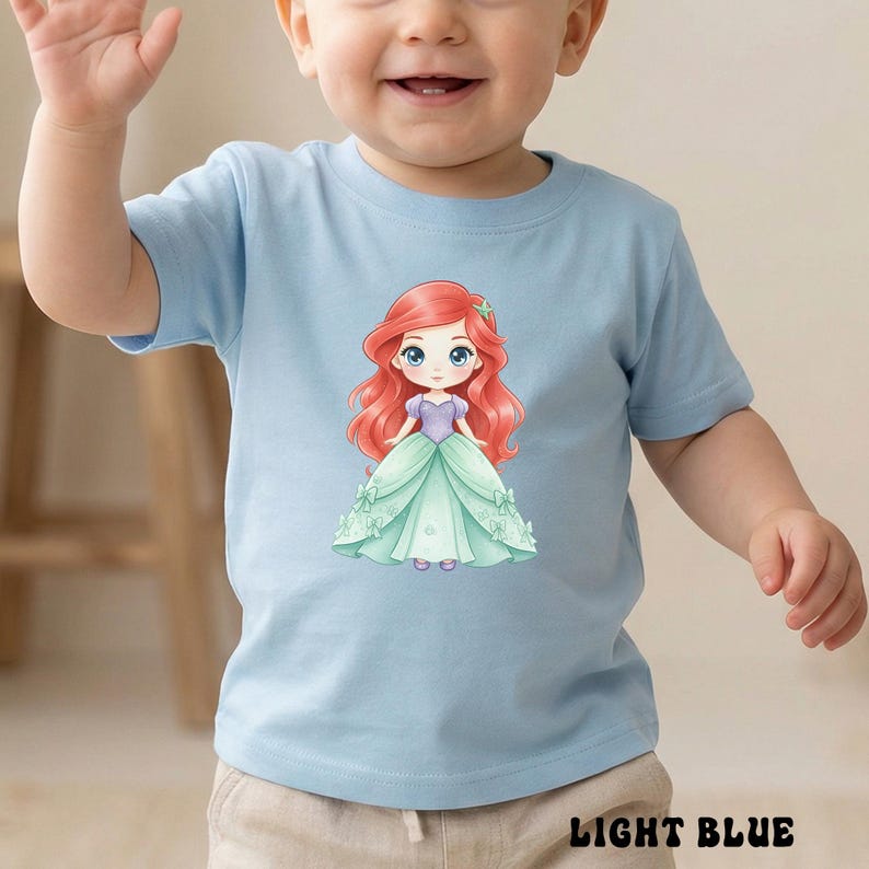 Puede incluir: Una camiseta de manga corta de color azul claro con un estampado gr&aacute;fico de un personaje de dibujos animados con pelo largo rojo, un corpi&ntilde;o morado y un vestido verde claro. El personaje tiene grandes ojos azules y un accesorio verde.