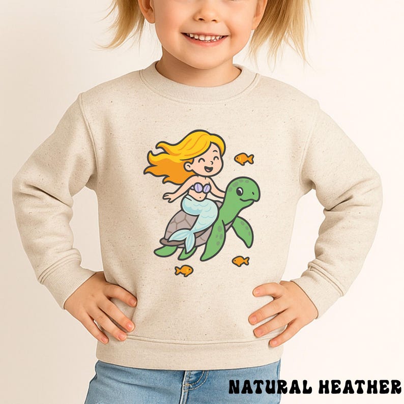 Puede incluir: Sudadera beige con una sirena de dibujos animados montada en una tortuga verde, rodeada de peces naranjas. La sirena tiene el pelo rubio y una cola azul. El texto "NATURAL HEATHER" est&aacute; impreso en la parte inferior.