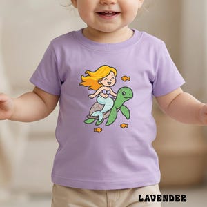 Puede incluir: Camiseta de color lavanda con una sirena de dibujos animados montada en una tortuga verde, con peces naranjas. La sirena tiene el pelo rubio y una cola azul. La palabra "LAVENDER" est&aacute; impresa en la parte inferior.