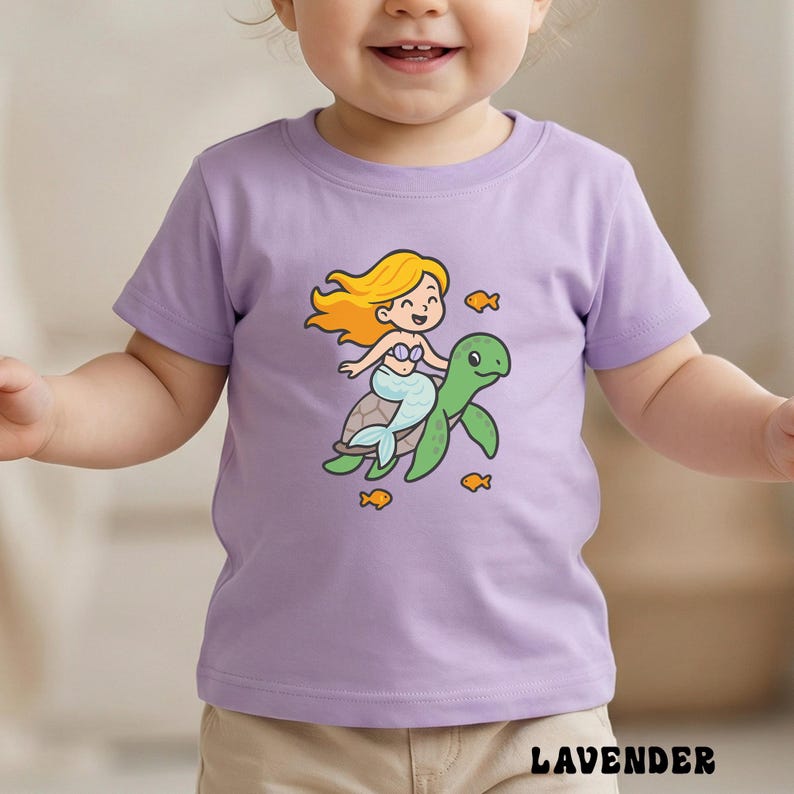 Puede incluir: Camiseta de color lavanda con una sirena de dibujos animados montada en una tortuga verde, con peces naranjas. La sirena tiene el pelo rubio y una cola azul. La palabra "LAVENDER" est&aacute; impresa en la parte inferior.