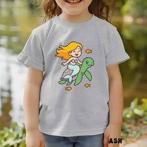 Puede incluir: Camiseta gris claro con una sirena de dibujos animados montada en una tortuga verde, rodeada de peces naranjas. La sirena tiene pelo rubio y una cola azul. La camiseta est&aacute; hecha de un material suave.